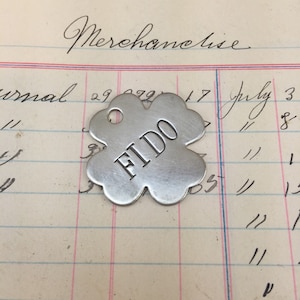 May include: A silver clover-shaped dog tag with the word "FIDO" engraved on it. The tag is lying on a piece of lined paper with handwritten text.