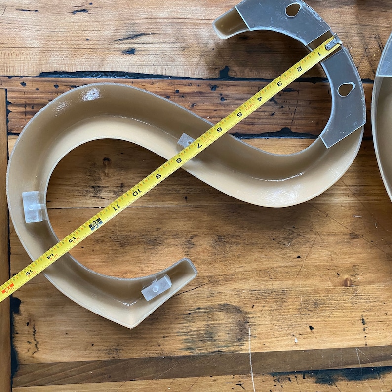 May include: A beige-colored, curved, S-shaped track with two white plastic pieces attached to the track. A yellow measuring tape is laid across the track, measuring 15 inches.