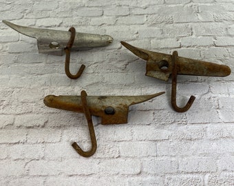 Peut inclure: Trois crochets en métal rouillé avec une extrémité pointue et un crochet à l'autre extrémité. Les crochets sont accrochés à un mur de briques blanches.