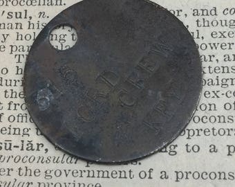 May include: A round, metal tag with the words "FORD CREW KPY" engraved on it. The tag is slightly worn and has a hole punched in the top.