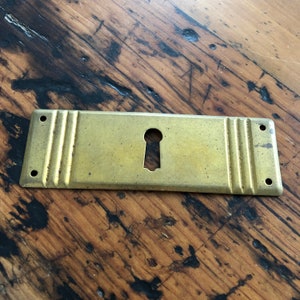 May include: A brass keyhole cover with a rectangular shape and three vertical lines on each side. The cover has a keyhole in the center and two holes on each end.