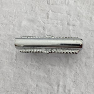 Puede incluir: Un tirador de cajón plateado con un borde decorativo texturizado. El tirador es de forma rectangular y tiene un acabado liso y pulido.