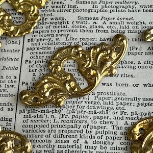 Pode incluir: Enfeites de metal dourado com intrincados desenhos florais e de rolagem. Estas peças de estilo vintage estão dispostas sobre um fundo de páginas de livros antigos, perfeitas para artesanato e joalheria.