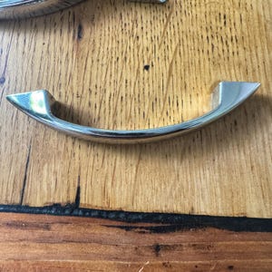 May include: A shiny, curved metal handle with a polished finish. The handle has a rounded center and angled ends, designed for pulling. The hardware is set against a wooden surface.