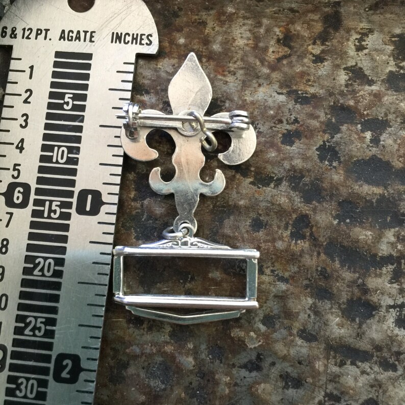 May include: Silver-toned pendant featuring a fleur-de-lis design with a rectangular frame hanging from the bottom. The pendant is approximately 3.8 cm tall and has a pin back.