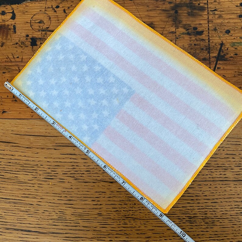 Pu&ograve; includere: Un patch di bandiera americana in tessuto bianco con un bordo giallo. Il patch &egrave; largo circa 23 cm e alto 15 cm.