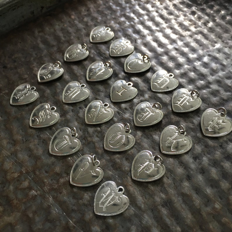 May include: A collection of silver heart-shaped charms, each with a different letter engraved on it. The charms are arranged in a pattern on a textured surface.