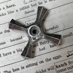 May include: A silver metal cross-shaped object with a round center hole. The object is lying on top of a page of a book with text about a hare, a rabbit, and a hen sitting on eggs.