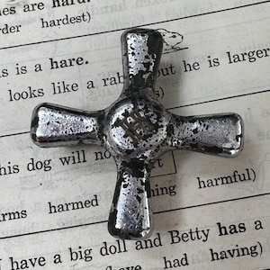 Puede incluir: Un objeto metálico en forma de cruz con un acabado plateado desgastado, sobre una página de texto impreso. El texto incluye las palabras "duro", "liebre" y "dañino". El objeto tiene cuatro brazos redondeados.