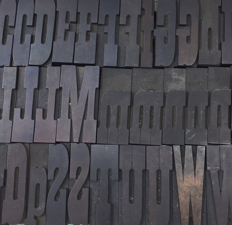 May include: A close-up of a collection of vintage wooden letterpress printing blocks. The blocks are arranged in rows and columns, and they feature the letters C, D, E, F, I, L, M, and O. The blocks are made of dark wood and have a worn, rustic appearance.