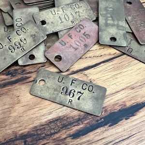 May include: Close-up of several brass tags with the text "U.F. CO." and a number followed by the letter "R". The tags are worn and have a rustic appearance.
