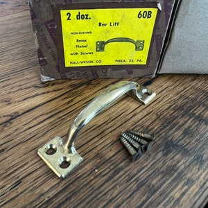 Puede incluir: Una caja de 2 docenas de manijas de elevación de barra de latón plateado no oxidables con tornillos. La caja está etiquetada con "60B Bar Lift" y "Hall-Wessel Co. Phila. 23, PA."