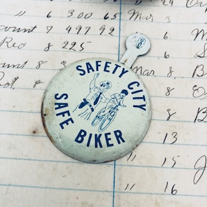 Könnte beinhalten: Ein weißer Knopf mit blauem Text und einer blauen Illustration eines Polizisten und eines Kindes auf einem Fahrrad. Der Text auf dem Knopf lautet "Safety City Safe Biker".