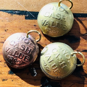 May include: Three gold-colored metal charms with the words "No Cash Value" engraved on them. The charms have a loop at the top for attaching to a necklace or bracelet.