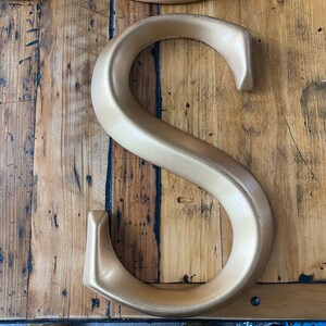 May include: A gold-colored metal letter S, with a smooth, curved surface. The letter is sitting on a wooden surface.