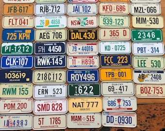 May include: A collection of vintage license plates from different states in the United States. The plates are arranged in rows and columns on a wooden surface. The plates are all different colors and have different designs. Some of the plates have the state name and year on them, while others have the state name and a slogan. The plates are a great way to add a touch of Americana to your home decor.