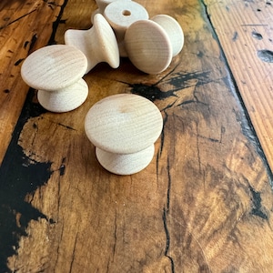Könnte beinhalten: Eine Gruppe von sechs unfertigen Holzgriffen mit runder Oberseite und zylindrischer Basis. Die Griffe sind in einer Gruppe auf einer Holzoberfläche angeordnet.