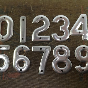 Puede incluir: Diez placas de números de metal plateado, cada una con un número diferente del 0 al 9. Los números están ligeramente en relieve y tienen un aspecto vintage.