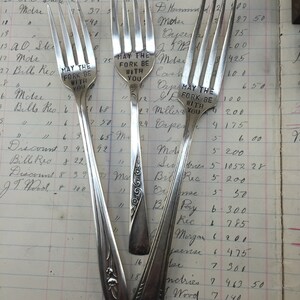May include: Three silver forks with engraved messages. The forks have ornate designs on the handles. The messages read "May the fork be with you".