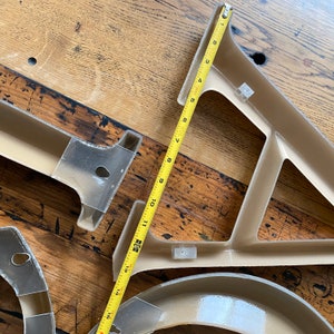 May include: A close-up of a beige plastic shelf bracket with a yellow measuring tape. The tape measures 15 inches. The shelf bracket is made of a lightweight plastic and has a simple design.