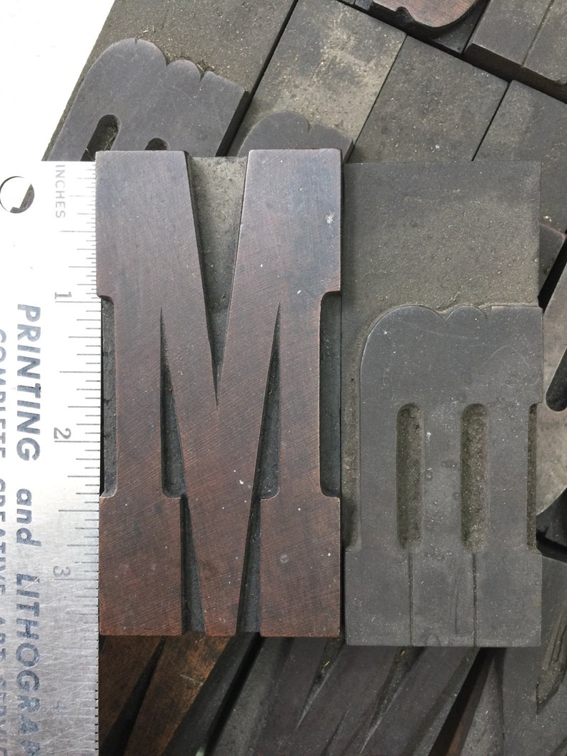 May include: Two wooden letterpress blocks, one with the letter "M" in a dark brown color and the other with the letter "m" in a gray color. The blocks are approximately 2 inches tall.