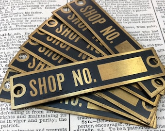 May include: A collection of vintage shop number signs. The rectangular signs are black with gold lettering and borders, featuring the text "SHOP NO." and a blank space. Each sign has two holes for mounting.