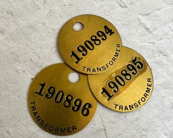 May include: Three round, gold-coloured metal tags with black text. Each tag has a hole in the centre. The text on the tags reads "190894 TRANSFORMER", "190895 TRANSFORMER", and "190896 TRANSFORMER".
