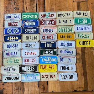 May include: A collection of vintage license plates from different states in the United States. The plates are arranged in rows and columns on a wooden surface. The plates are all different colors and have different designs. Some of the plates have state names and slogans, while others have numbers and letters.