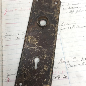 May include: A vintage brass door plate with a keyhole and a round hole in the center. The plate is rectangular and has a worn, aged look.