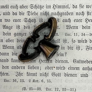 May include: A close-up of a small, antique, brass bell with a decorative, ornate, metal handle. The bell is sitting on a page of a book with German text. The text reads "Wenn ab das Schalk ist, so wird d in dir ist, Fir sein?"