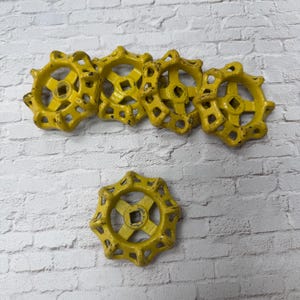 May include: A collection of six yellow, star-shaped valve handles with a distressed finish. The handles have a central square opening and are arranged on a white brick background. The handles are likely made of metal.