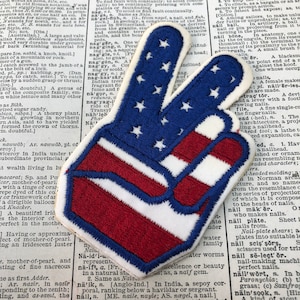 May include: A peace sign patch with the American flag design. The patch is embroidered with red, white, and blue thread. The patch is on a white background.