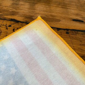 Pu&ograve; includere: Primo piano di un angolo di un tessuto bianco con un bordo giallo. Il tessuto presenta un sottile motivo a righe in tonalit&agrave; di rosa, blu e giallo.