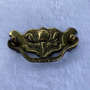 Może przedstawiać: Złoty metalowy uchwyt do szuflady z kwiatowym wzorem. Uchwyt ma zakrzywiony uchwyt i dwa okrągłe pokrętła.