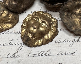 May include: Close-up of several antique brass lion head embellishments. The lion heads are detailed with manes and facial features. The metal has a tarnished, aged appearance, suggesting vintage or repurposed use. The background is a vintage paper with handwritten text.
