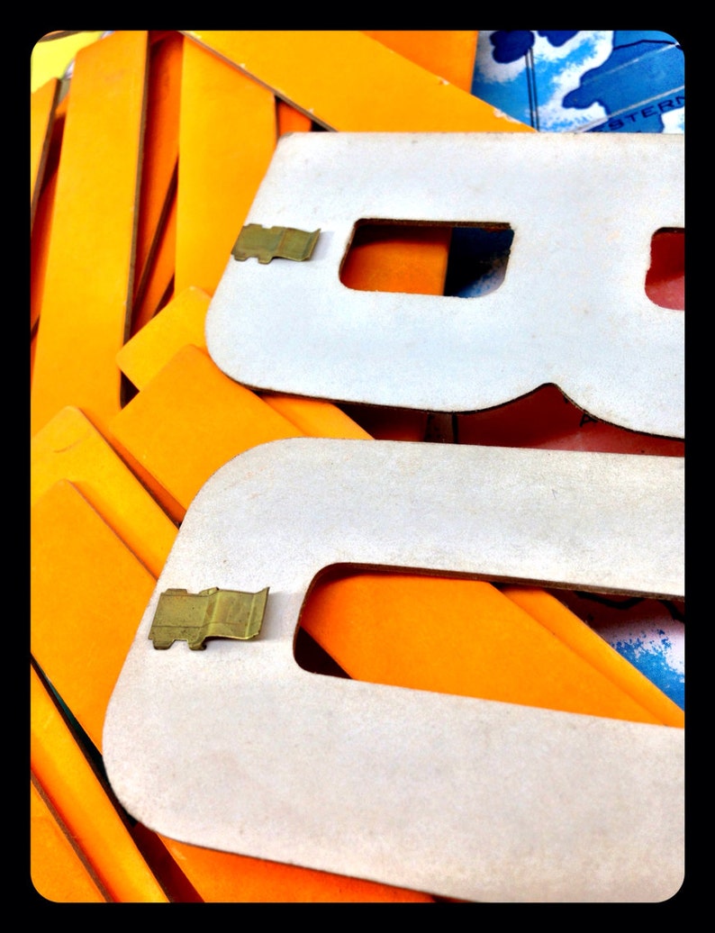 May include: White cardboard letters with metal clips, forming the letter "B". The letters are stacked on top of orange cardboard.