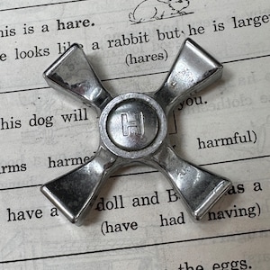 May include: A silver metal four-pronged object with a letter H in the center. The object is lying on a page of a book with text about hares, dogs, and hens.