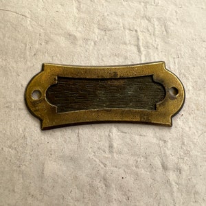 May include: A brass nameplate with a rectangular opening for engraving. The plate has a slightly curved shape and a decorative border.