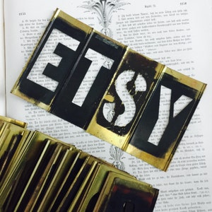 以下が含まれることがあります： 「ETSY」という単語を綴る、真鍮製のレターステンシル4枚セット。ステンシルは黒と金で、ビンテージな外観です。