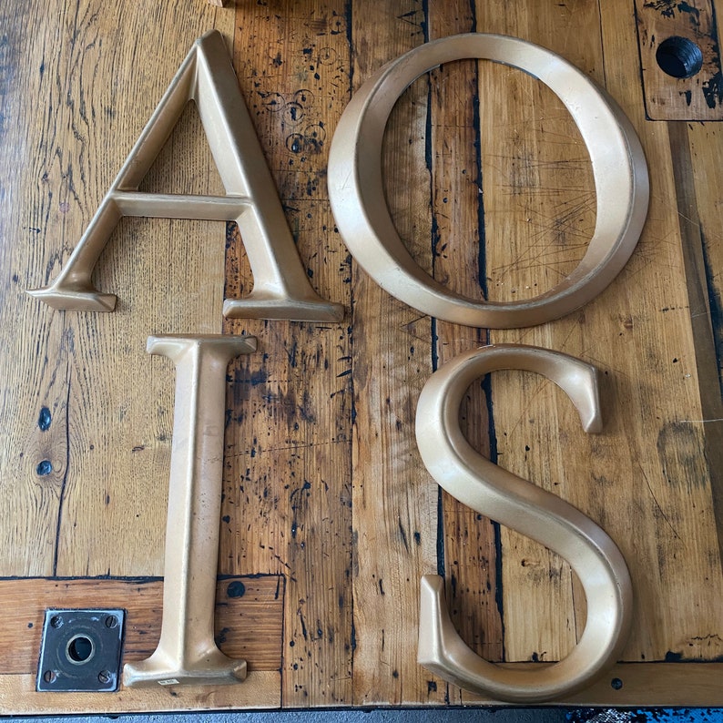 May include: Gold-painted wooden letters spelling out the word "AIOIS" on a wood background.