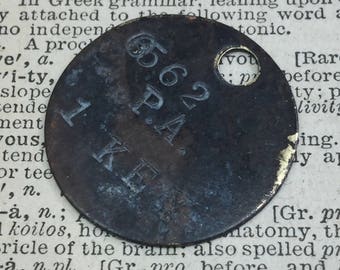 May include: A round, dark metal tag with the numbers "65.62" and the letters "P.A." and "KE" stamped on it. The tag has a hole in the centre.