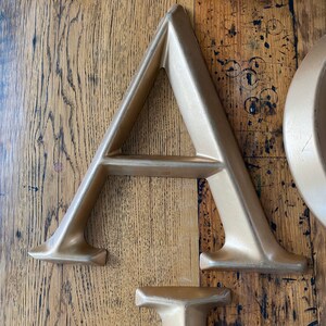 May include: A gold-colored metal letter A with a shelf in the middle. The letter is sitting on a wooden surface.