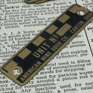 May include: A brass tag with black lettering that reads "UNIT NO. AMERICAN MINE DOOR CO. CANTON, OHIO". The tag has a round hole in the top right corner and four rectangular raised areas.