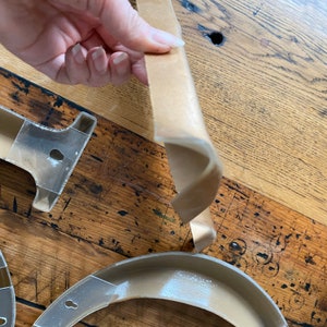 May include: A close-up of a brown paper tape being held by a hand. The tape is curved and has a slight sheen. The tape is on a wooden surface.