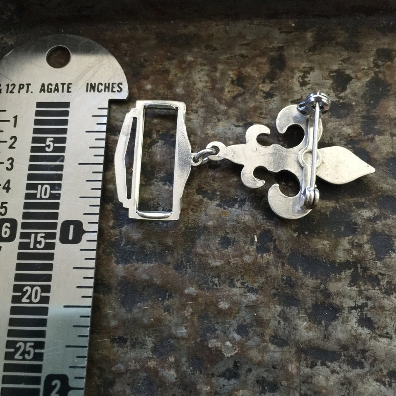 May include: Silver-toned pendant featuring a fleur-de-lis design with a chain and a clasp. The pendant is approximately 3.8 cm in length.