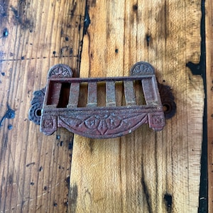 May include: A rusty cast iron wall mounted rack with a decorative floral design and five slots. The rack is painted a reddish brown color.