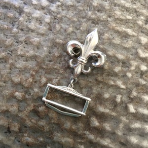 May include: Silver-toned metal belt buckle featuring a fleur-de-lis design and a rectangular frame.