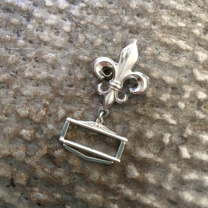 May include: Silver-toned metal belt buckle featuring a fleur-de-lis design and a rectangular frame.