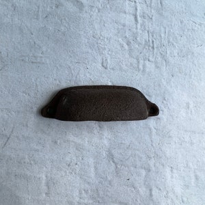 May include: A dark brown cast iron drawer pull with a slightly curved shape. The pull has a rough, textured surface.