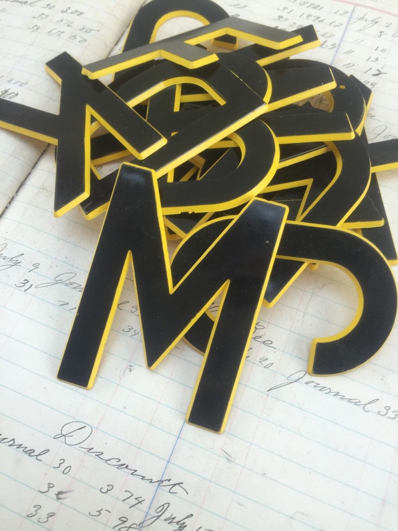 May include: A pile of black plastic letters with yellow outlines. The letters are in various sizes and are stacked on top of each other. The letters are on a white and yellow lined surface.